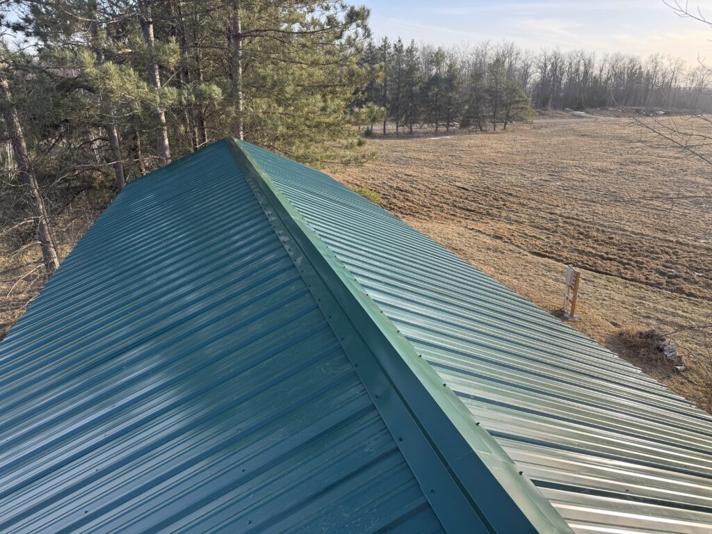 A green metal roof viewed from above, surrounded by trees on the left and an open, grassy field on the right under a clear sky.