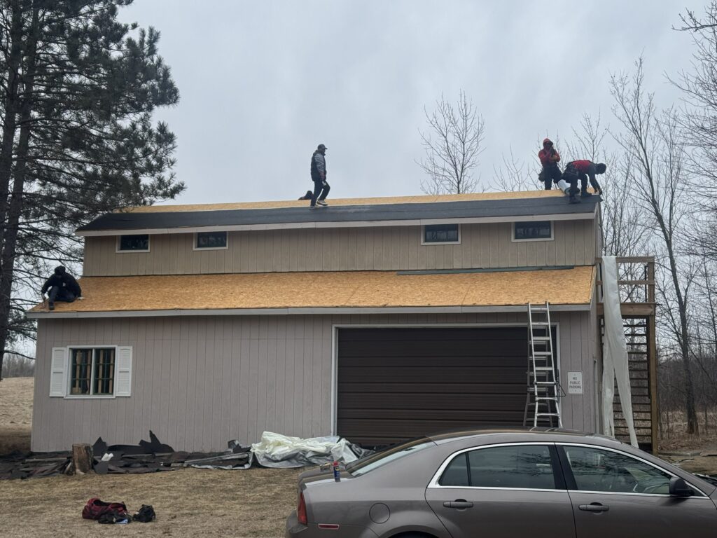 Four people are working on the roof of a two-story garage or barn, installing new roofing material. A car is parked in front, and ladders lean against the building. Leafless trees and overcast skies are in the background.