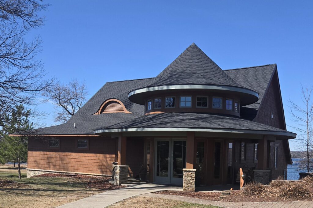 A modern house with a unique conical roof, large windows, and stone pillars. It is surrounded by leafless trees, with a lake visible in the background under a clear blue sky.