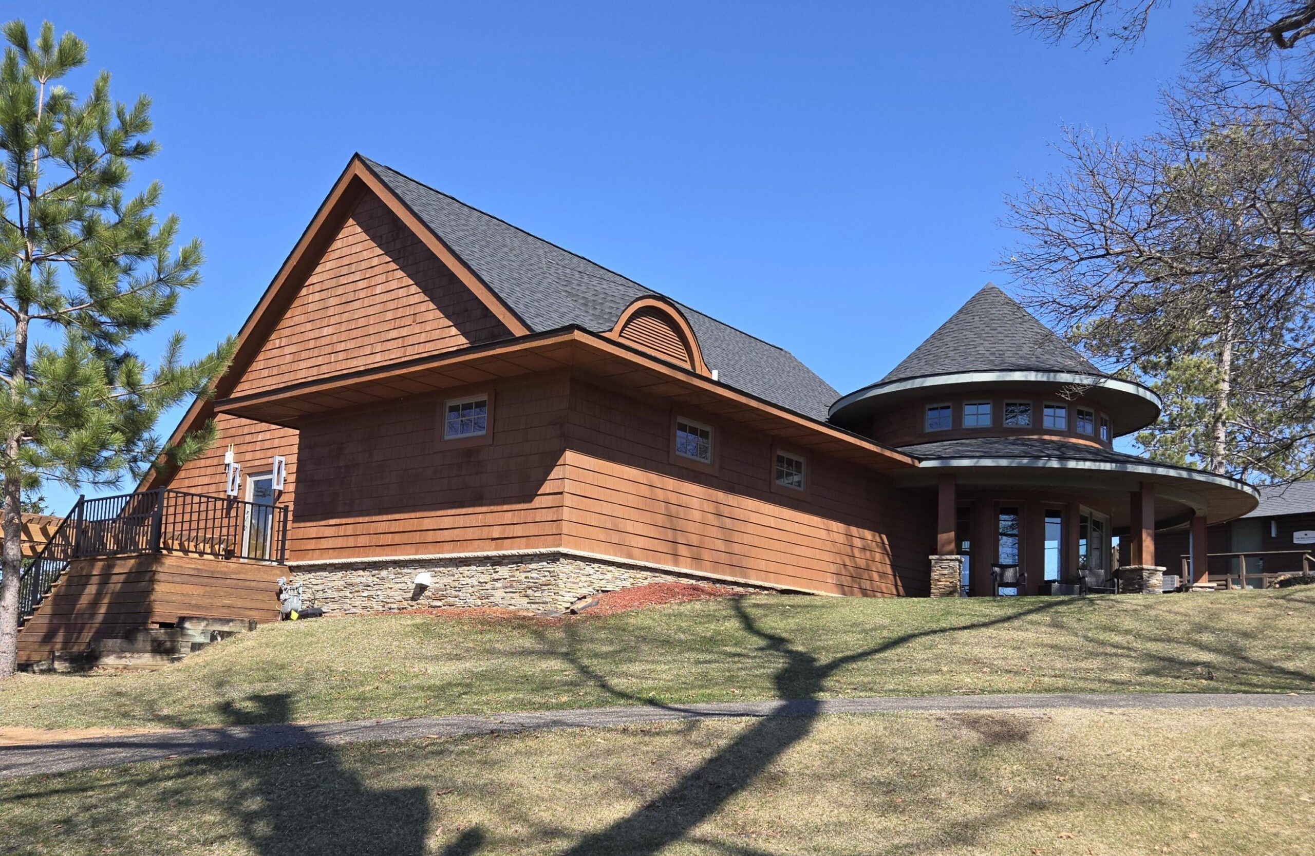 A large house with reddish-brown siding and a round turret, featuring a dark gray roof, stone foundation, and a grassy front lawn under a clear blue sky.