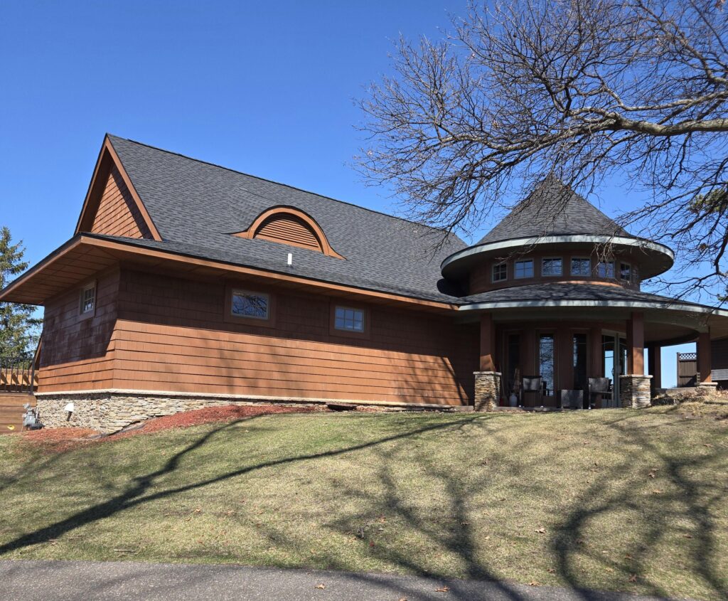 A unique house with brown siding, stone trim, and a turret-like round section with large windows, sits on a grassy lawn under a clear blue sky. Leafless tree branches cast shadows on the ground.
