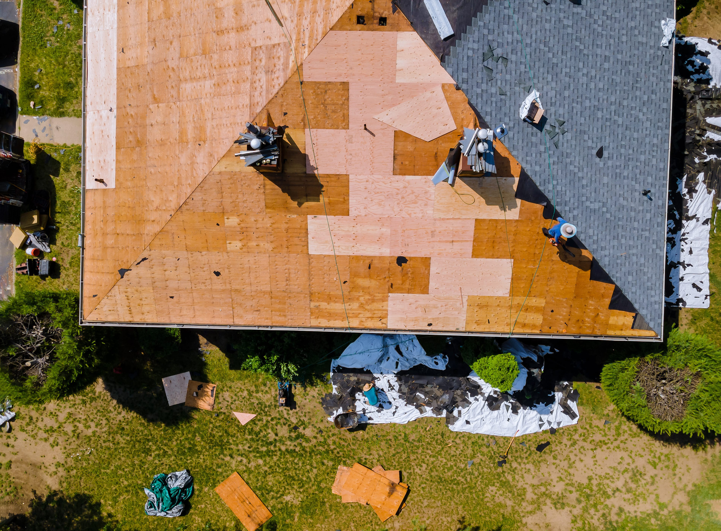 Aerial view of a house undergoing roof repairs; half of the roof is covered in new plywood, while the other half has old shingles. Various tools, materials, and tarps are scattered on the ground below.