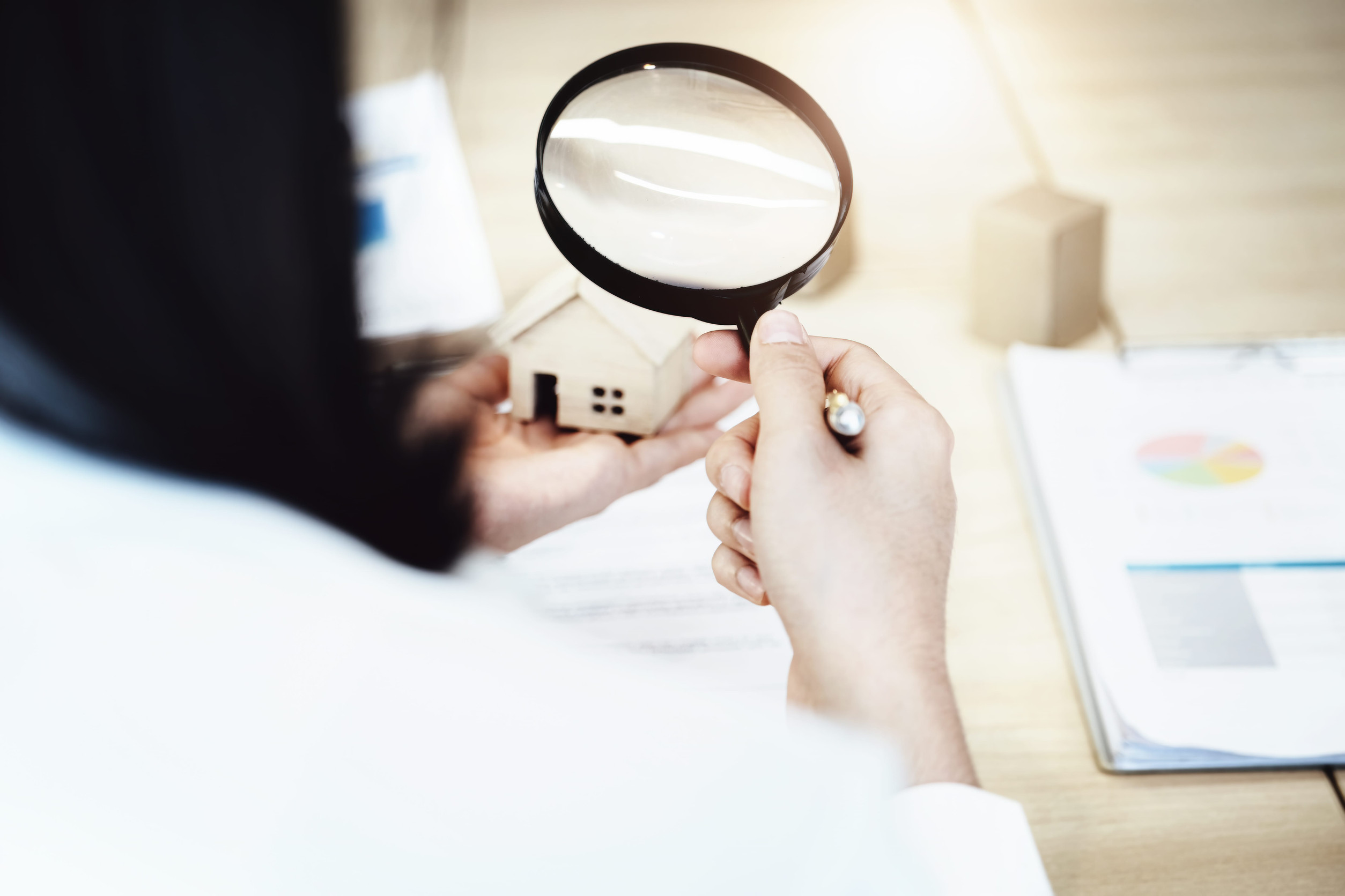 Person holding a magnifying glass over a small model house, with papers and a pen nearby on a desk, suggesting close examination or analysis of real estate or financial documents.
